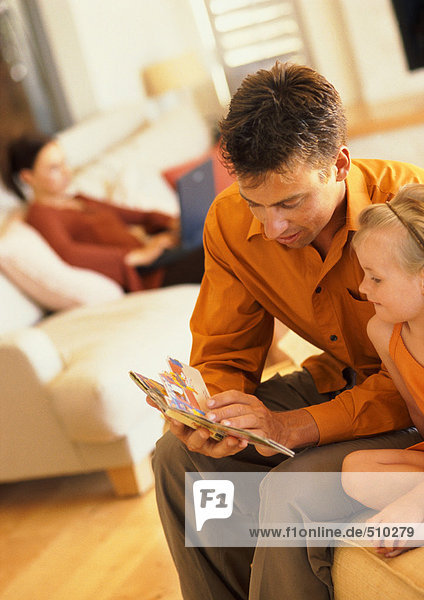 Father and daughter looking at book  woman sitting on sofa in background