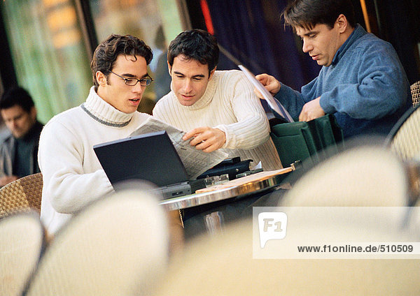 Drei Männer sitzen auf der Terrasse des Cafés und sehen sich Dokumente an.