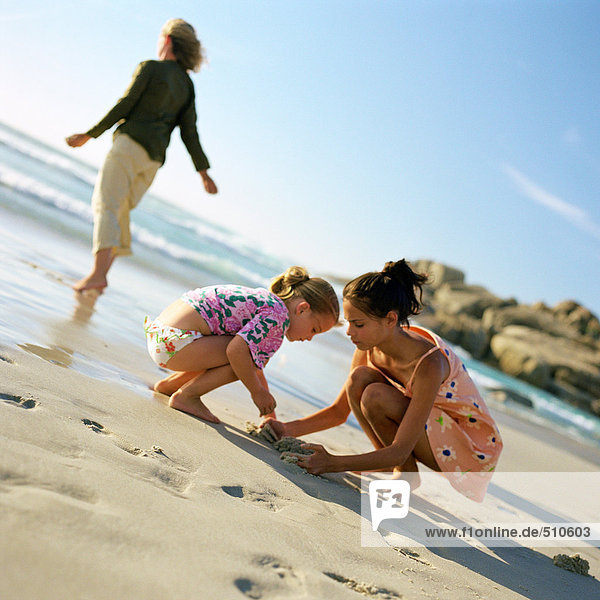Two girls playing in sand while mother looks at the sea