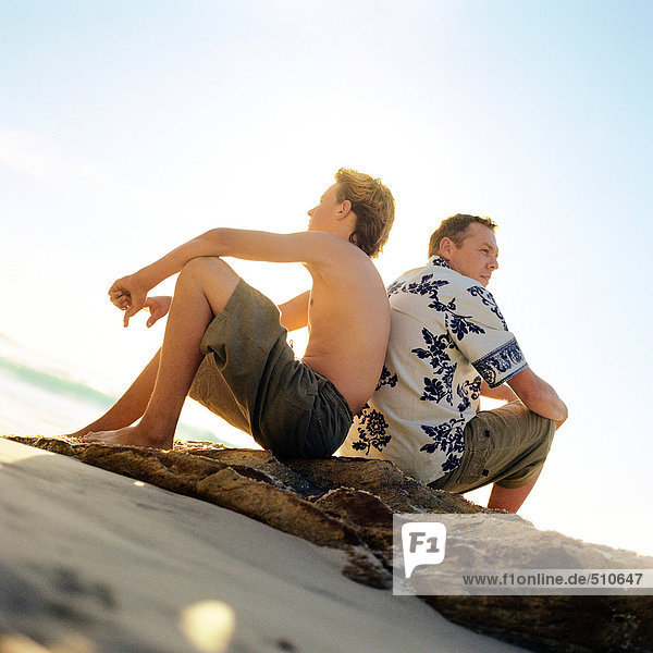 Mann und Junge sitzen Rücken an Rücken am Strand.
