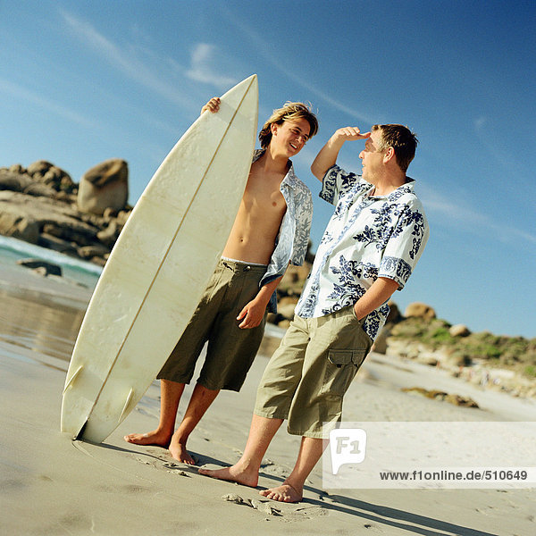 Mann steht neben dem Jungen mit Surfbrett am Strand