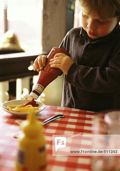 Kleiner Junge hinter dem Tisch mit rot-weiß karierter Tischdecke  Ketchup auf Pommes frites drücken