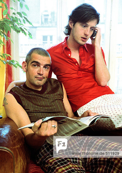 Zwei Männer sitzen  einer schaut in die Kamera  hält Zeitung  einer telefoniert mit dem Handy.