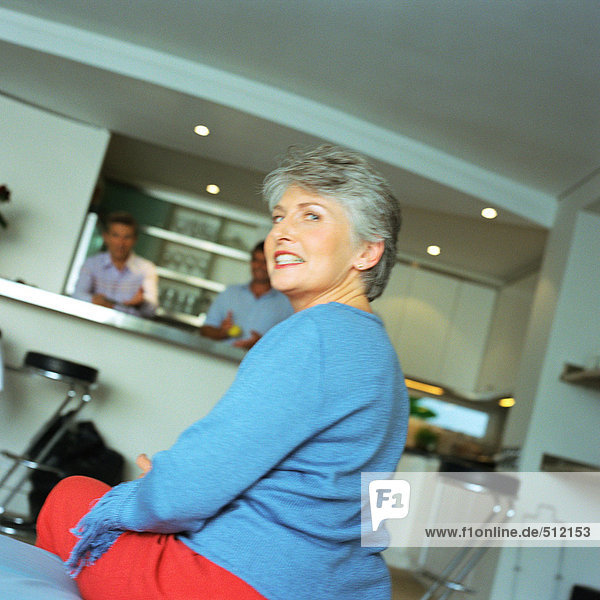 Mature woman sitting  looking over shoulder  men standing in kitchen in background