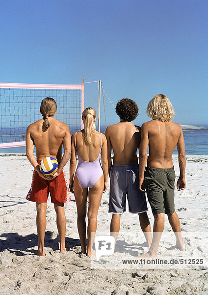 Vier junge Leute stehen am Strand mit Volleyball  Rückansicht.