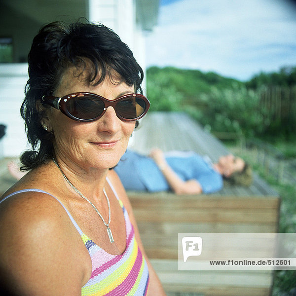 Reife Frau auf einer Bank sitzend  mit Sonnenbrille  zweite Person im Hintergrund liegend