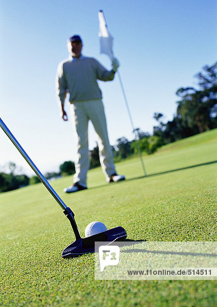 Golfer mit Fahne auf Grün  Putter und Ball im Vordergrund