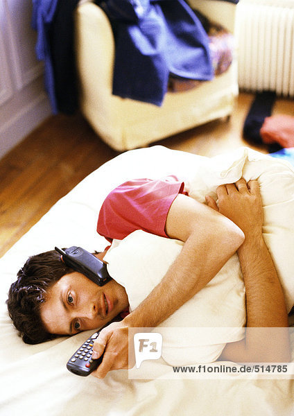 Mann auf dem Bett liegend mit Telefon und Fernbedienung