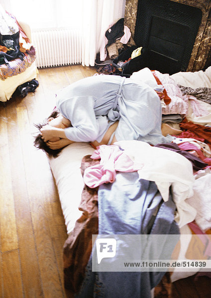 Woman huddled up on bed among clothes with hands on head