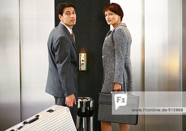 Geschäftsmann und Geschäftsfrau warten auf den Aufzug und schauen über die Schultern in die Kamera.
