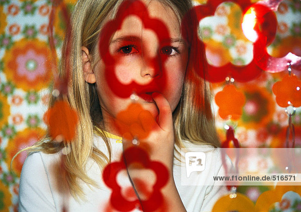 Kleines Mädchen mit Finger im Mund  das durch eine hängende Blumengirlande schaut.