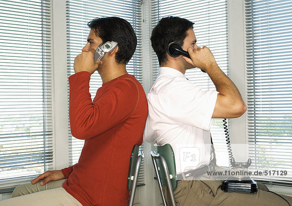 Zwei Männer sitzen Rücken an Rücken  einer mit Handy  der andere mit Drehtelefon.