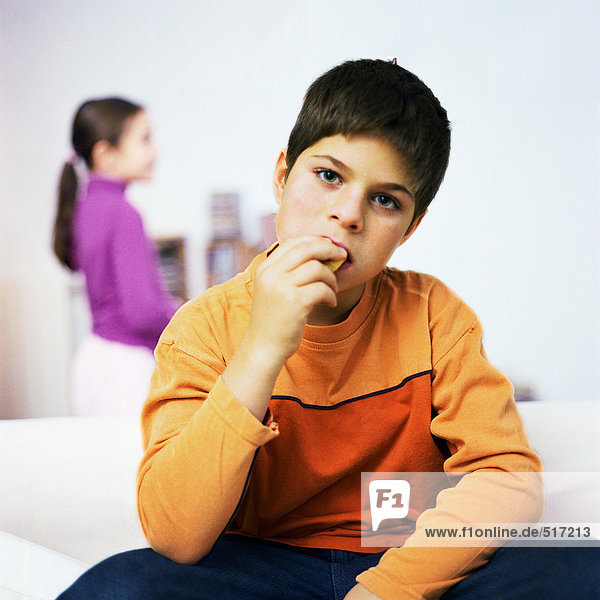 Junge sitzt auf dem Sofa  isst  Mädchen steht im Hintergrund
