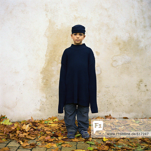 Junge steht zwischen Herbstblättern und trägt einen übergroßen Pullover.
