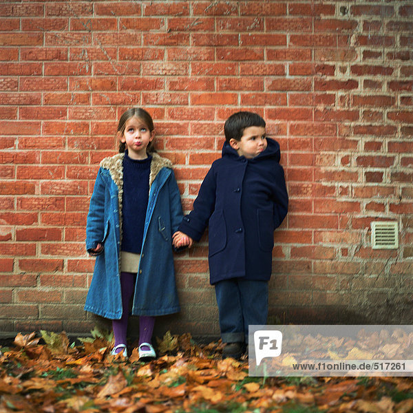 Boy and girl holding hands and making faces in front of brick wall  full length