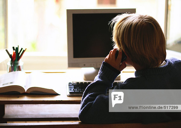 Young boy sitting at computer talking on phone  rear view