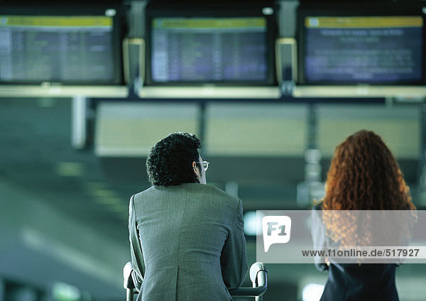Geschäftsmann und Geschäftsfrau unter den Abflug- und Ankunftsmonitoren des Flughafens  Rückansicht