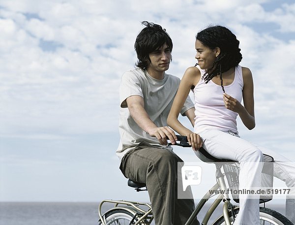 Junger Mann sitzt auf dem Fahrrad  junge Frau sitzt auf dem Fahrradkorb und schaut über die Schulter auf den jungen Mann.