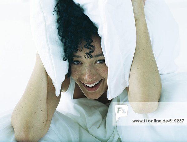 Frau im Bett  die Kissen zu beiden Seiten des Kopfes hält  mit Blick auf die Kamera