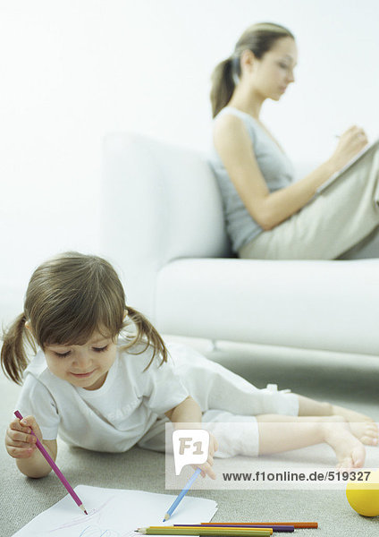 Kleines Mädchen auf dem Boden liegend  junge Frau sitzt hinter ihr auf der Couch und schreibt mit den Knien nach oben.