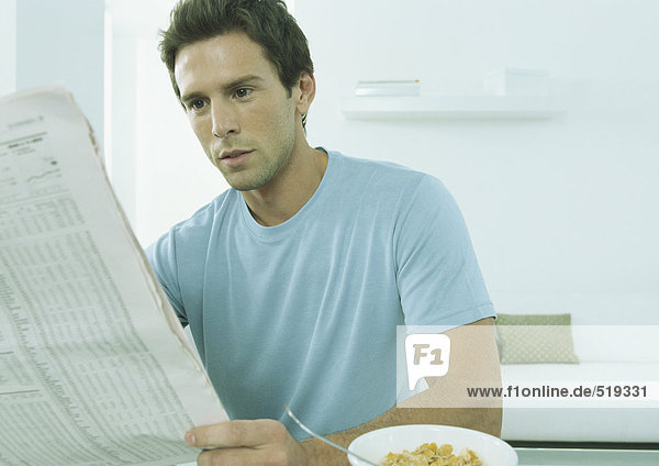 Junger Mann sitzt am Tisch mit Getreideschale und hält Finanzseite der Zeitung