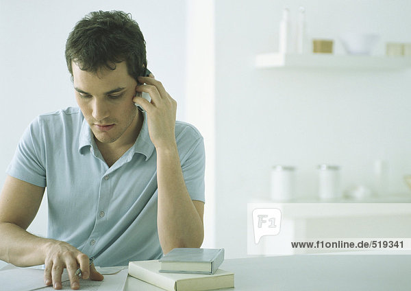 Junger Mann im Polohemd am Tisch sitzend mit Büchern und Papieren  Handy haltend  nach unten schauend