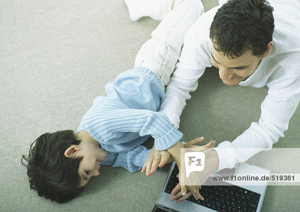 Mann und kleiner Junge liegen auf dem Boden und spielen mit dem Laptop.