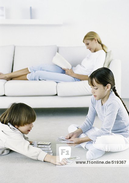 Mädchen und Junge spielen Karten auf dem Boden,  Frau sitzt auf dem Sofa und liest im Hintergrund.