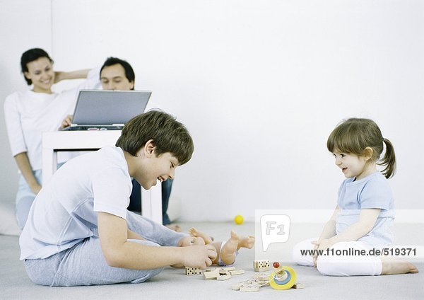Boy and girl sitting on floor playing  man and woman using laptop in background