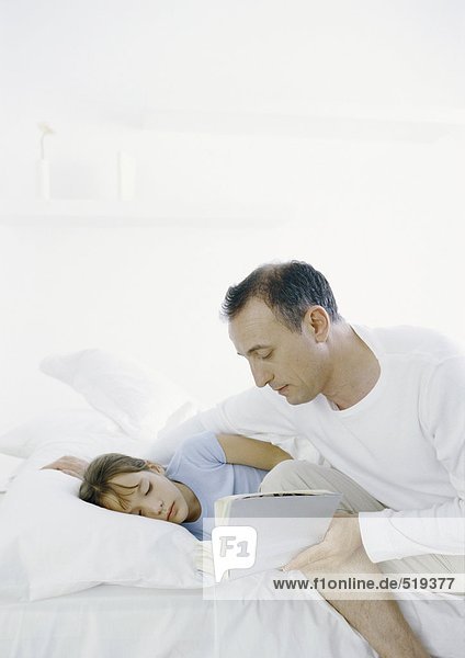 Man sitting on edge of bed reading book to girl lying in bed