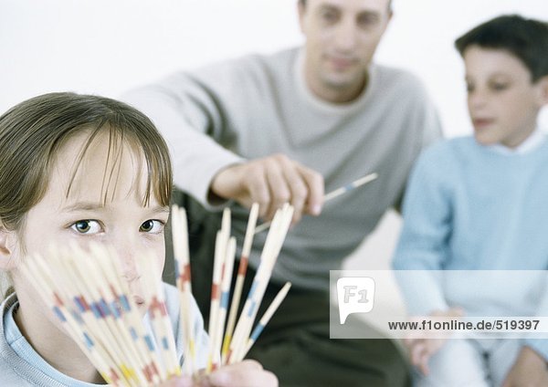 Girl holding out pick up sticks  father and boy in background