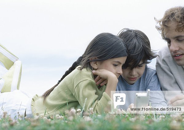 Junge und Mädchen mit Vater auf Mägen auf Gras liegend  mit Blick auf Handy