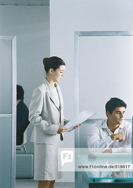 Businesswoman holding document  standing next to man sitting at keyboard in office