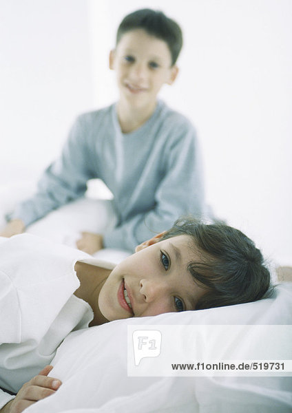 Mädchen im Bett liegend mit Blick auf die Kamera  Junge im Hintergrund