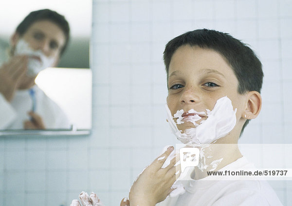 Junge mit Rasiercreme auf Gesicht und Händen  Mann rasiert im Hintergrund