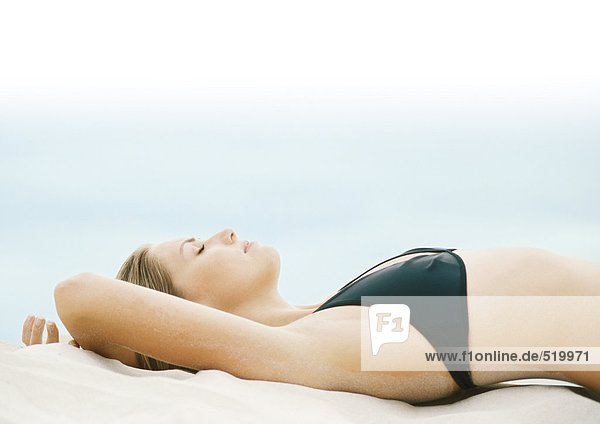 Junge Frau im Bikini auf Sand liegend mit geschlossenen Augen  Seitenansicht