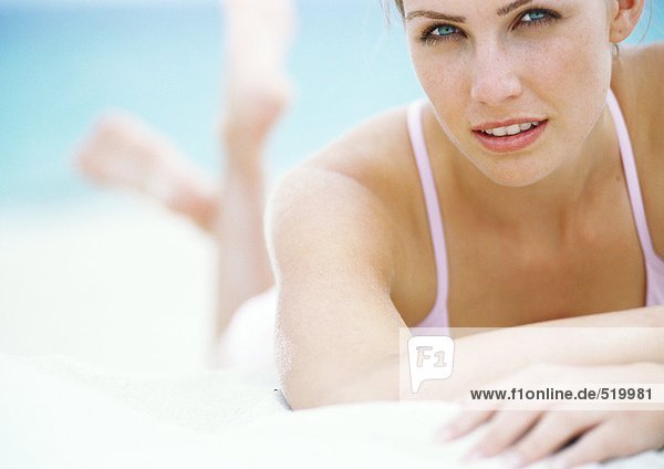 Junge Frau im Badeanzug auf dem Bauch im Sand liegend  mit Blick auf die Kamera