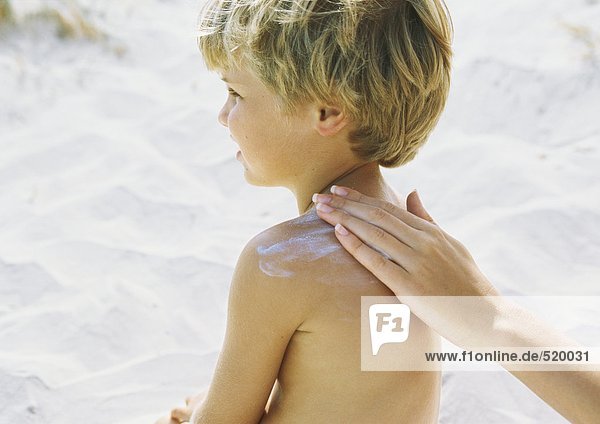 Kleiner Junge am Strand mit Sonnencreme von Mutter in die Schulter gerieben