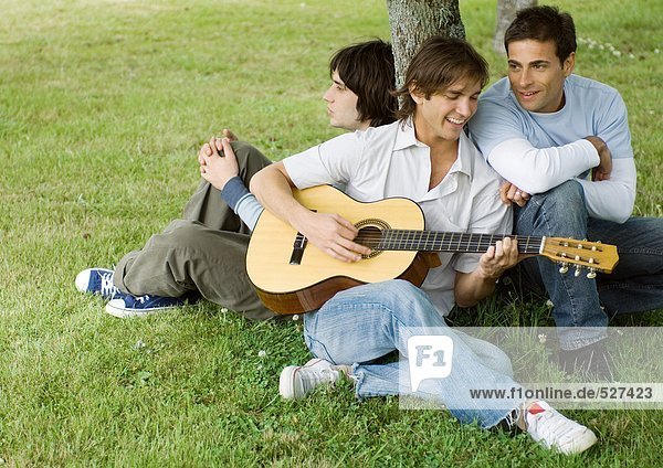 Young male friends sitting near tree  playing guitar