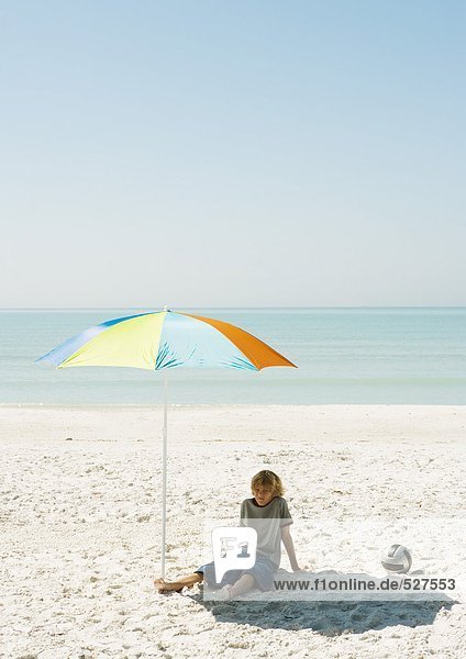 Junge am Strand unter dem Sonnenschirm sitzend