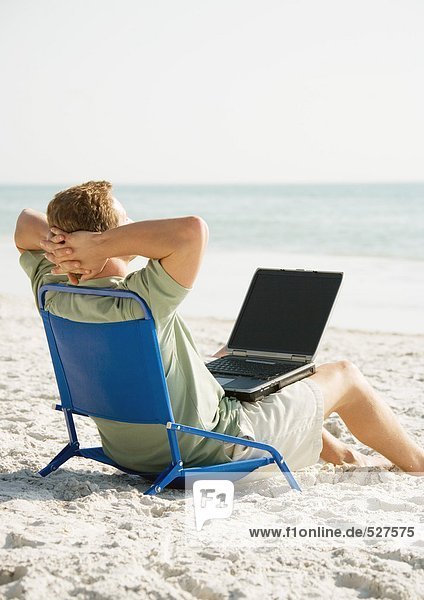 Mann am Strand sitzend mit Computer auf dem Schoß