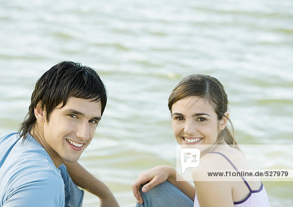 Young couple sitting side by body of water  smiling and looking at camera