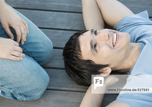 Young man reclining on the ground with hands behind head  smiling  someone kneeling beside him  partial view