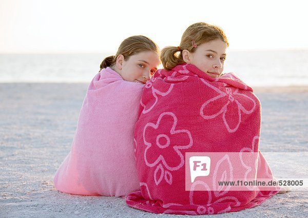 Zwei Mädchen sitzen in Handtücher gewickelt am Strand.