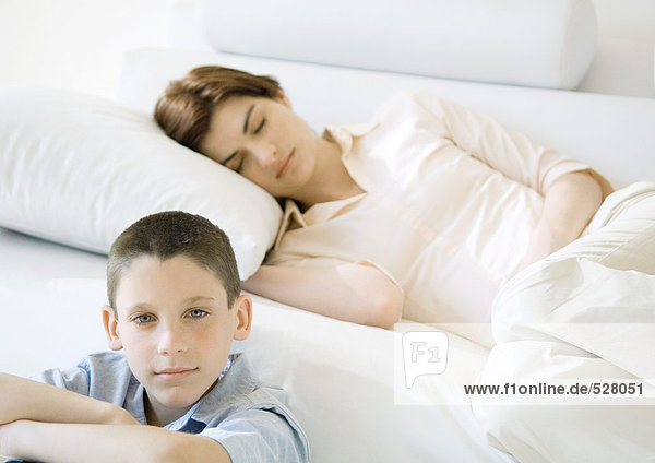 Boy sitting near sofa where mother is napping