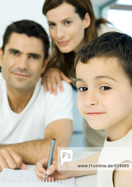 Junge macht Hausaufgaben mit Eltern im Hintergrund