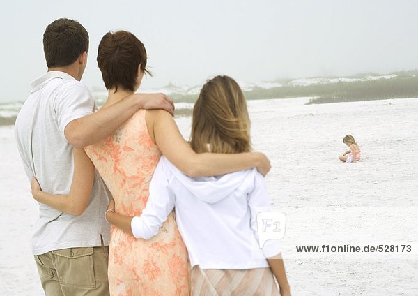 Parents and teenage daughter watching little girl play in sand in distance