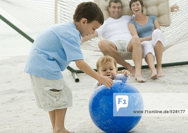 Junge und Baby spielen mit dem Ball  während die Eltern von der Hängematte aus zusehen.