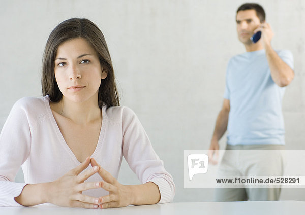 Woman sitting  looking at camera while man uses cell phone in background