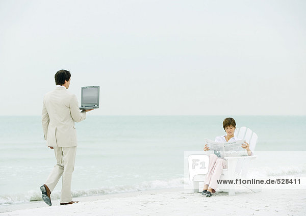 Frau sitzt am Strand und liest Zeitung als Geschäftsmann mit offenem Laptop